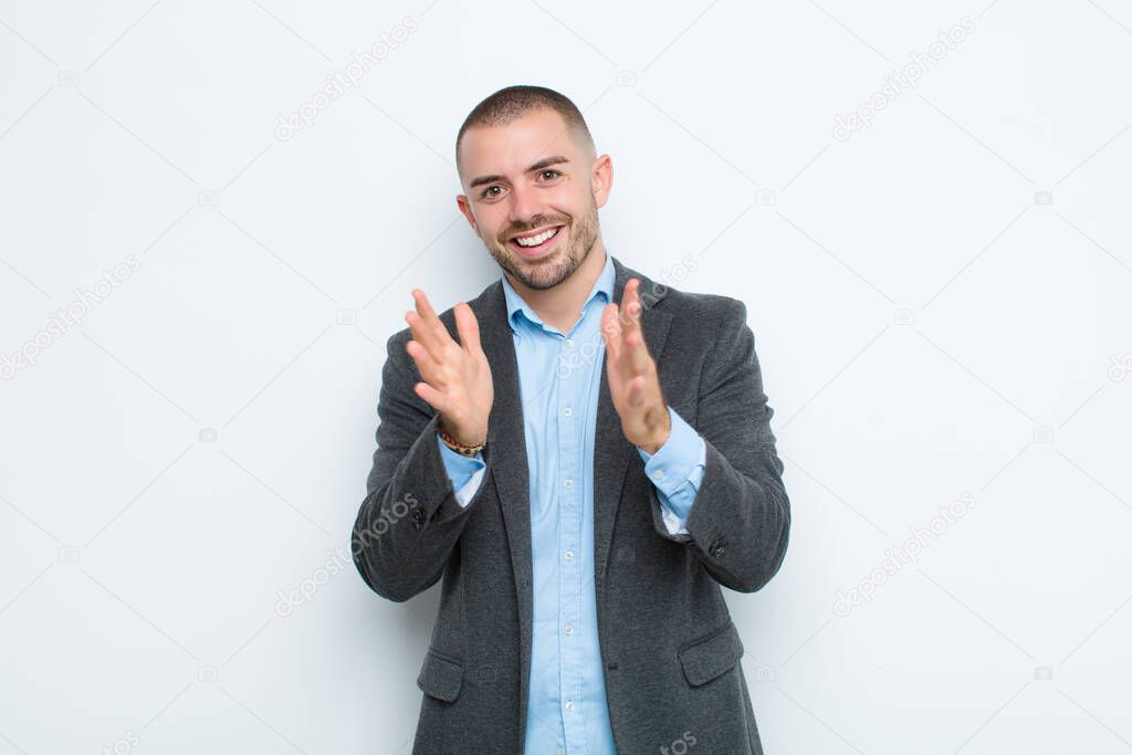 young businessman feeling happy and successful, smiling and clapping hands, saying congratulations with an applause against flat wall