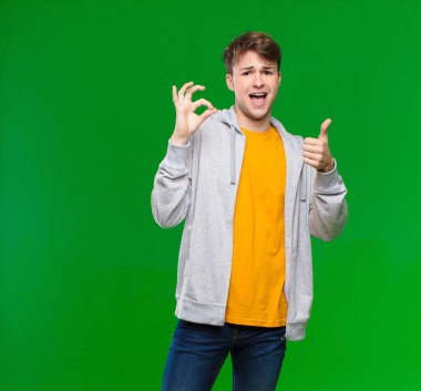 Genç sarışın adam mutlu, şaşkın, tatmin olmuş ve şaşırmış hissediyor, tamam gösteriyor ve parmaklarını havaya kaldırıyor, kroma tuşuna gülümsüyor.
