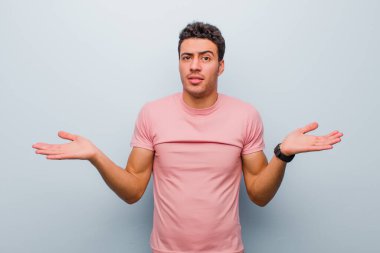 Genç Arap adam şaşkın, kafası karışmış ve stresli görünüyor, farklı seçenekler arasında merak ediyor, gri duvara karşı kararsız hissediyor.
