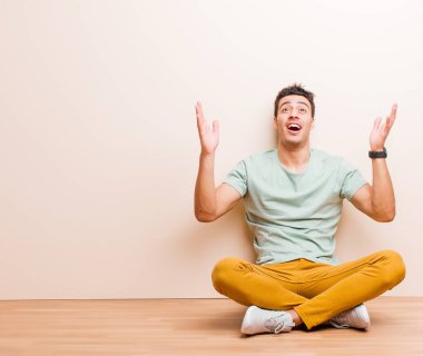 Genç Arap adam kendini mutlu, şaşkın, şanslı ve şaşırmış hissediyor. Zaferi kutlarken iki eli havada yerde oturuyor.