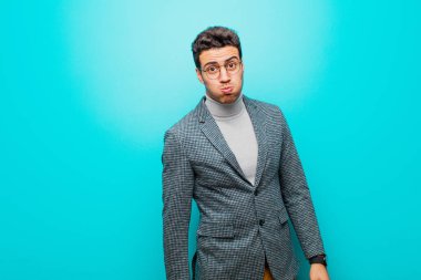 Genç Arap adam şapşal, çılgın, şaşırmış bir ifadeyle, yanakları şişmiş, doldurulmuş, şişman ve mavi duvarda yemek dolu.