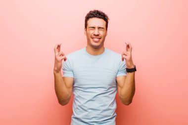 Genç Arap adam gülümsüyor ve endişeyle iki parmağını çapraz yapıyor. Endişeli ve pembe duvara karşı iyi şanslar diliyor.