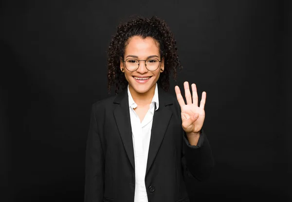 young black woman smiling and looking friendly, showing number four or fourth with hand forward, counting down against black wall