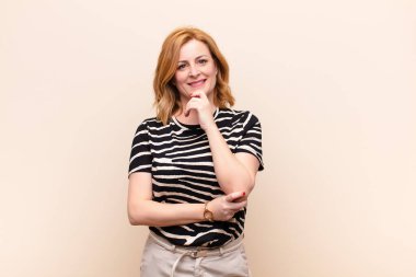 Orta yaşlı kadın mutlu görünüyor ve gülümsüyor. Elleri çenesinde, merak ediyor ya da soru soruyor. Seçenekleri karşılaştırıyor.