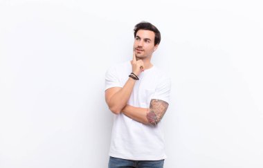 Genç yakışıklı adam mutlu bir şekilde gülümsüyor ve hayal kuruyor ya da şüphe ediyor, beyaz duvara bakıyor.