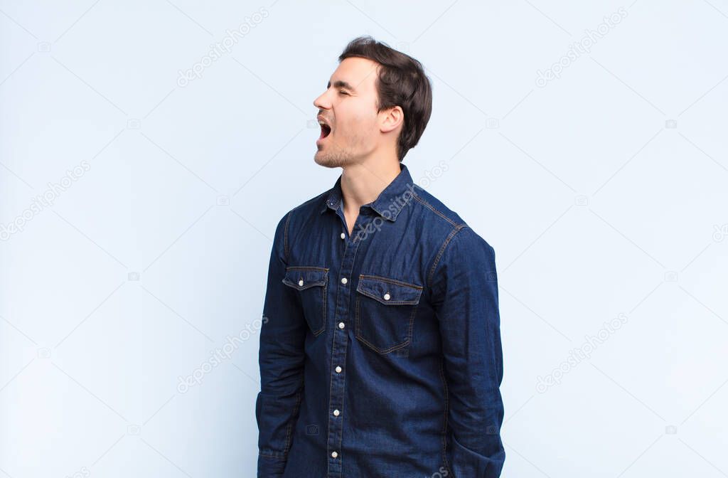 young handsome man screaming furiously, shouting aggressively, looking stressed and angry against blue wall