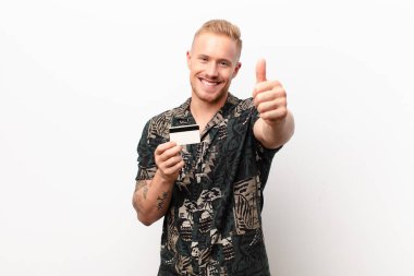 Genç sarışın adam gururlu, kaygısız, kendinden emin ve mutlu, kredi kartıyla olumlu gülümsüyor.
