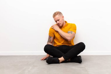 Genç sarışın adam yorgun, stresli, endişeli, hayal kırıklığına uğramış ve depresyonda, sırtı ya da boynu ağrıyan çimento zemininde oturuyor.