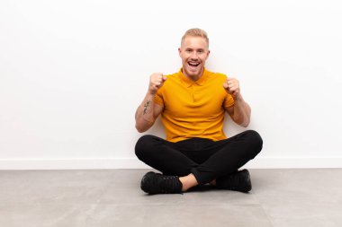 Genç sarışın adam kendini mutlu, şaşırmış ve gururlu hissediyor. Bağırıyor ve başarıyı kutluyor. Büyük bir gülümsemeyle çimentoda oturuyor.