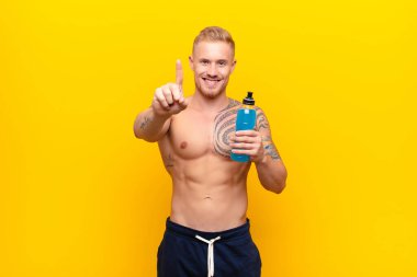 Genç sarışın güçlü adam gururla ve kendinden emin bir şekilde gülümsüyor. Bir numaralı pozunu başarıyla veriyor. Enerji içeceğiyle lider gibi hissediyor.