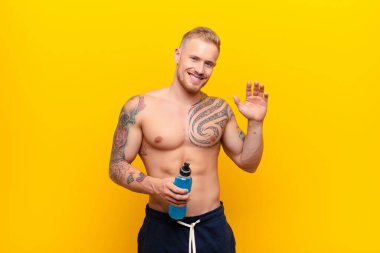 Genç sarışın güçlü adam mutlu ve neşeli bir şekilde gülümsüyor, el sallıyor, seni selamlıyor, ya da enerji içeceğiyle veda ediyor.