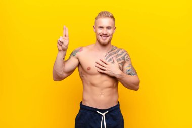 Genç güçlü sarışın adam mutlu, kendinden emin ve güvenilir görünüyor, gülümsüyor ve zafer işareti gösteriyor, sarı duvara karşı pozitif bir tutum sergiliyor.