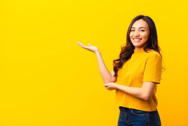 Genç Latin güzel kadın gururla ve güvenle gülümsüyor, mutlu ve tatmin olmuş hissediyor ve düz duvara karşı fotokopi alanında bir konsept gösteriyor.