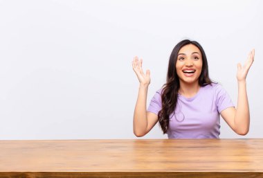 Genç, Latin, güzel bir kadın kendini mutlu, şaşkın, şanslı ve şaşırmış hissediyor. İki eli havada, düz duvara karşı zaferi kutluyor.