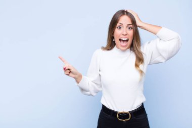 Genç ve güzel İspanyol kadın gülüyor, mutlu, pozitif ve şaşırmış görünüyor, mavi duvara karşı yanal fotokopi alanını işaret eden harika bir fikrin farkına varıyor.