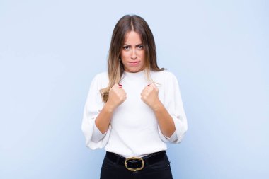 Genç ve güzel İspanyol bir kadın kendinden emin, sinirli, güçlü ve agresif görünüyor. Yumrukları mavi duvara karşı boks pozisyonunda dövüşmeye hazır.