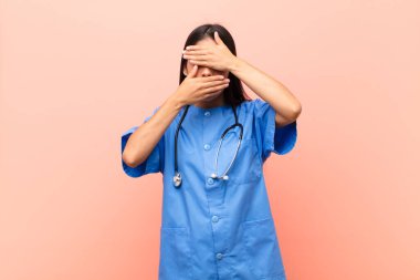 Genç Latin hemşire iki eliyle kameraya hayır diyerek yüzünü örtüyor! Resimleri reddetmek ya da pembe duvara yaslanan fotoğrafları yasaklamak