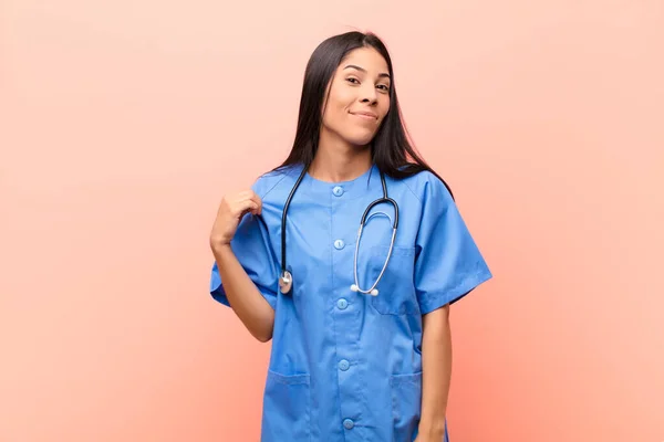 young latin nurse looking arrogant, successful, positive and proud, pointing to self against pink wall