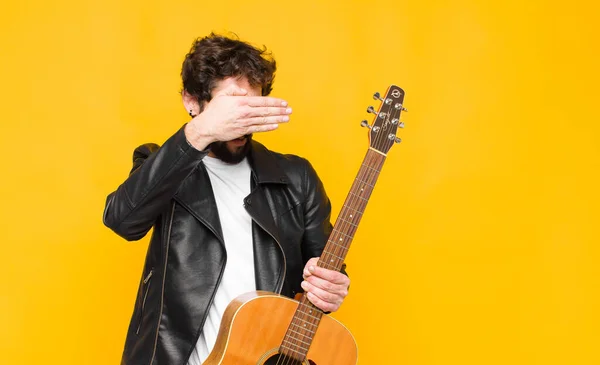 young musician man covering face with both hands saying no to the camera! refusing pictures or forbidding photos with a guitar, rock and roll concept