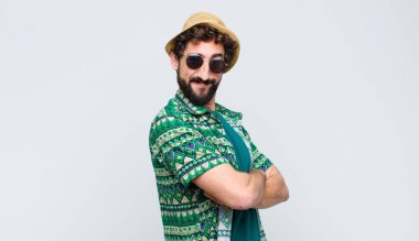 Kameraya çapraz kollarıyla gülümseyen genç turist ve mutlu, kendinden emin, tatmin olmuş bir ifade, beyaz duvara karşı yan bakış.