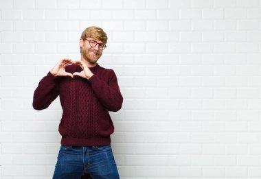 Genç sarışın adam gülümsüyor ve mutlu, sevimli, romantik ve aşık, iki eliyle kalbi şekillendiriyor.