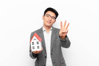Genç Japon adam gülümsüyor ve arkadaş canlısı görünüyor, üç ya da üçüncüyü gösteriyor, bir ev modeliyle geriye doğru sayıyor.