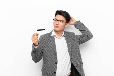 Genç Japon adam şaşkın ve şaşkın hissediyor, başını kaşıyor ve yana bakıyor. Kredi kartı kavramı.