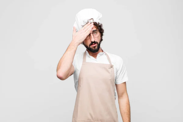 young crazy chef looking stressed, tired and frustrated, drying sweat off forehead, feeling hopeless and exhausted against white wall