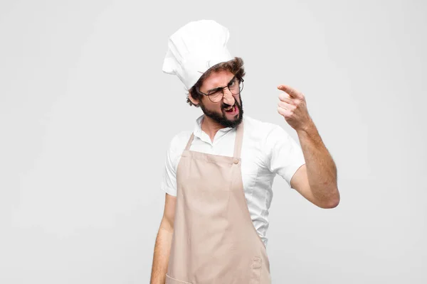 young crazy chef pointing at camera with an angry aggressive expression looking like a furious, crazy boss against white wall