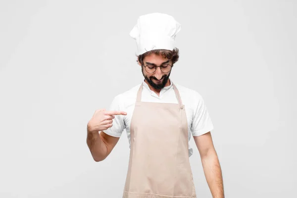 young crazy chef smiling cheerfully and casually, looking downwards and pointing to chest against white wall