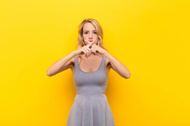 Genç sarışın kadın ciddi ve hoşnutsuz görünüyor. Reddedilmek için iki parmağını öne uzatmış, turuncu duvara karşı sessizlik istiyor.