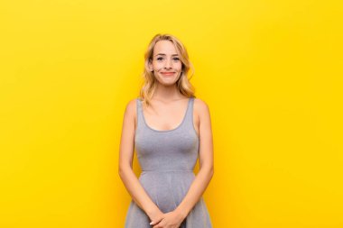 Genç sarışın kadın pozitif ve kendinden emin gülümsüyor. Tatmin olmuş, arkadaş canlısı ve turuncu duvarın karşısında mutlu görünüyor.