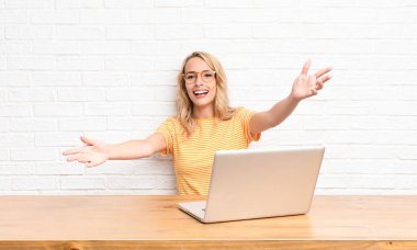 Genç sarışın kadın neşeli bir şekilde gülümsüyor sıcak, dostça, sevgi dolu bir kucaklama yapıyor, dizüstü bilgisayar kullanarak kendini mutlu ve sevimli hissediyor.