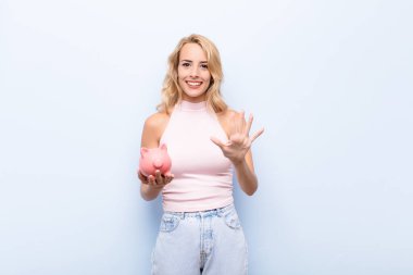 Genç sarışın kadın gülümsüyor ve arkadaş canlısı görünüyor, 5 ya da 5 numarayı gösteriyor, elinde kumbarayla geriye doğru sayıyor.