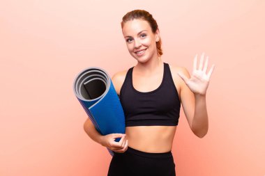 Kızıl saçlı güzel bir kadın gülümsüyor ve arkadaş canlısı görünüyor, 5 ya da 5 numarayı gösteriyor, elinde yoga minderiyle geriye doğru sayıyor..