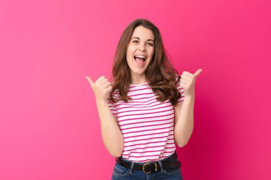 Genç, güzel bir kadın neşeyle gülümsüyor ve mutlu görünüyor. Kaygısız ve pozitif hissediyor. İki başparmağı da pembe duvarda.