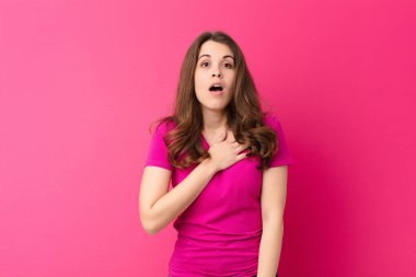 Genç ve güzel bir kadın şok olmuş ve şaşırmış hissediyor, gülümsüyor, el ele tutuşuyor, o kişi olmaktan mutlu ya da pembe duvara karşı minnettarlık gösteriyor.