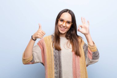 Genç, güzel bir kadın kendini mutlu, şaşkın, tatmin olmuş ve şaşırmış hissediyor. İyi olduğunu gösteriyor ve işaret ediyor. Mavi duvara gülümsüyor.