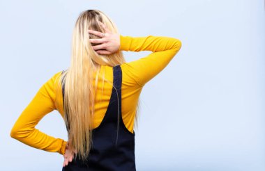 Genç, güzel sarışın kadın düşünüyor ya da şüphe ediyor, kafasını kaşıyor, kafası karışık ve kafası karışık hissediyor, düz duvara ya da arkaya bakıyor.