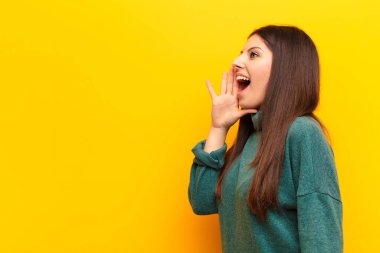 Genç ve güzel bir kadın profili, mutlu ve heyecanlı görünüyor, bağırıyor ve sarı duvara karşı alanı kopyalamak için çağırıyor.