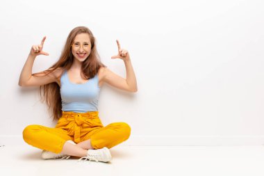 Genç sarışın kadın, çerçeveletiyor ya da kendi gülüşünü çiziyor iki eliyle, olumlu ve mutlu görünüyor, sağlık konsepti yerde oturuyor.