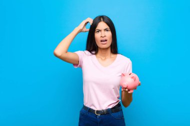 Genç, güzel, Latin bir kadın kafası karışmış ve kafası karışmış hissediyor. Kafasını kaşıyor ve düz duvara bakıyor.