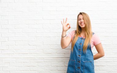 Genç sarışın kadın kendini mutlu, rahat ve tatmin olmuş hissediyor. Onayını iyi bir hareketle gösteriyor. Tuğla duvara gülümsüyor.