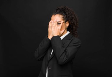 Genç siyahi kadın elleriyle yüzünü kapatıyor, parmaklarının arasından şaşkın bir ifadeyle bakıyor ve siyah duvara bakıyor.