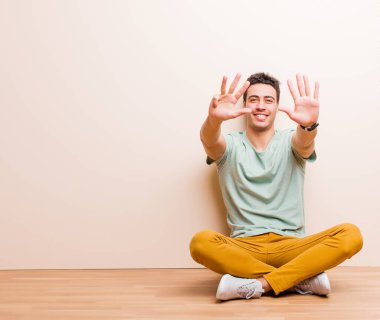 Genç Arap adam gülümsüyor ve dost canlısı görünüyor. Dokuz ya da dokuzuncu numarayı gösteriyor. Elleri önde, yerde oturmuş geri sayıyor.