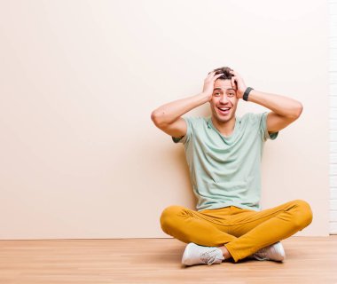 Genç Arap adam el ele tutuşuyor, ağzı açık, son derece şanslı, şaşırmış, heyecanlı ve mutlu bir şekilde yerde oturuyor.