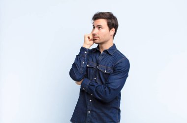 Yoğunlaşmış bakışlı genç yakışıklı adam, şüpheli bir ifadeyle merak ediyor, yukarı ve yana mavi duvara bakıyor