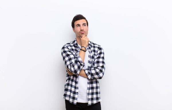 young handsome man thinking, feeling doubtful and confused, with different options, wondering which decision to make against white wall