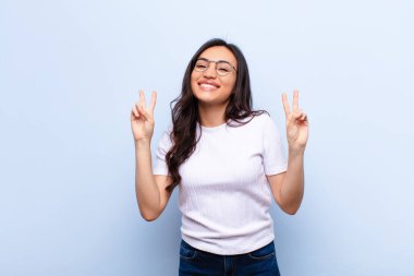 Genç Latin güzel kadın gülümsüyor ve mutlu görünüyor, arkadaş canlısı ve memnun, iki eli düz duvara karşı zafer ya da barış işareti yapıyor