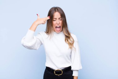 Genç ve güzel İspanyol kadın mutsuz ve stresli görünüyor, intihar jesti elinde silah işareti yapıyor, kafasını mavi duvara doğrultuyor.